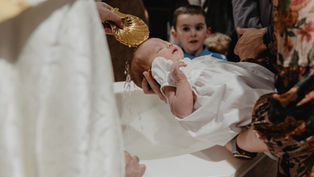 Priest pours water from a gold shell over a sleeping baby in a white gown during a baptism, with family watching nearby.