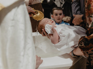 Priest pours water from a gold shell over a sleeping baby in a white gown during a baptism, with family watching nearby.