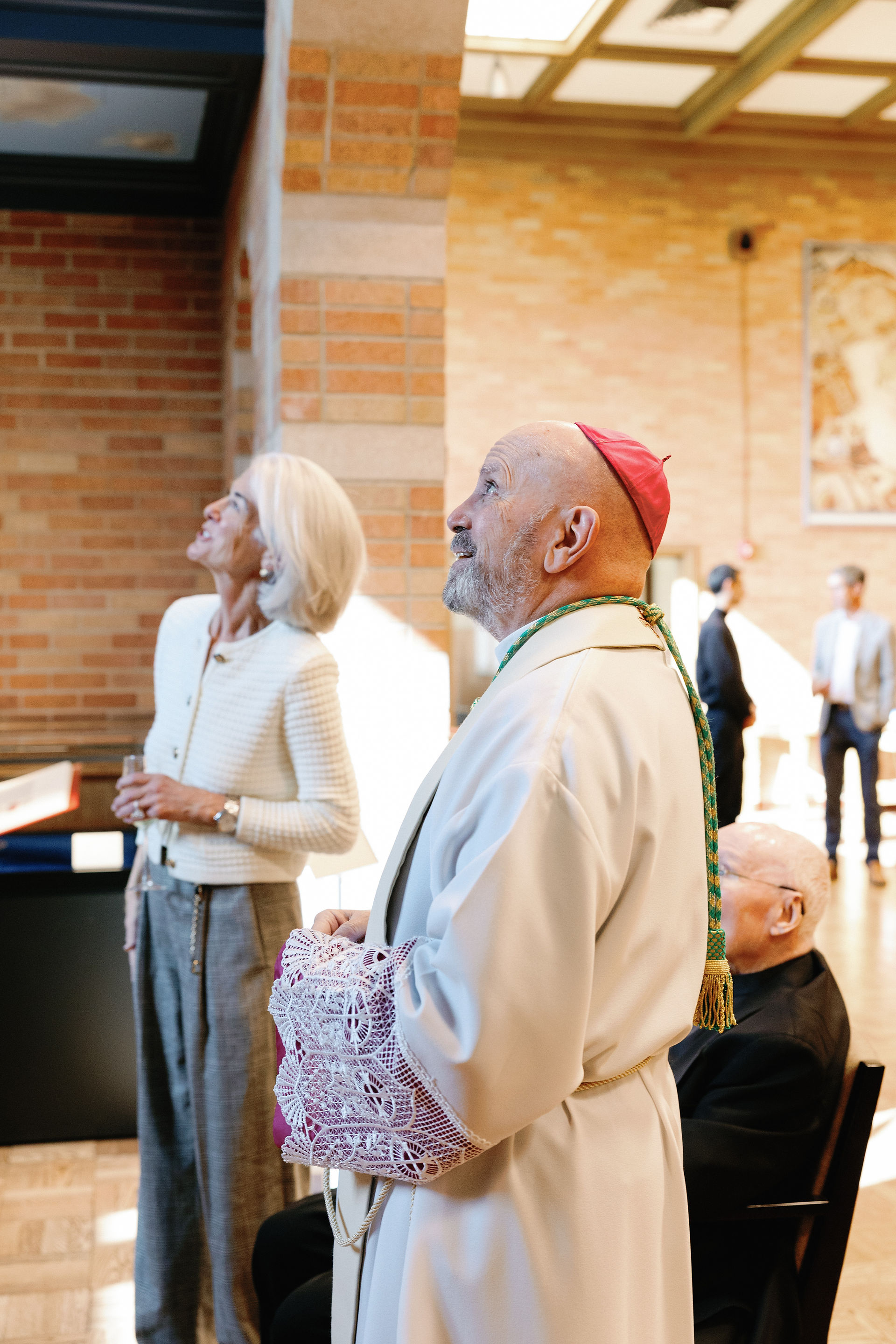 A clergy member and a woman in white look upward in a sunlit brick hall. Others are visible in the background, creating a focused atmosphere.