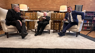 Three men sit on a stage in a library, engaged in conversation. Each holds a book or notebook. Warm lighting adds a cozy ambiance.