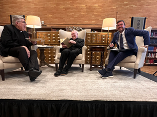 Three men sit on a stage in a library, engaged in conversation. Each holds a book or notebook. Warm lighting adds a cozy ambiance.