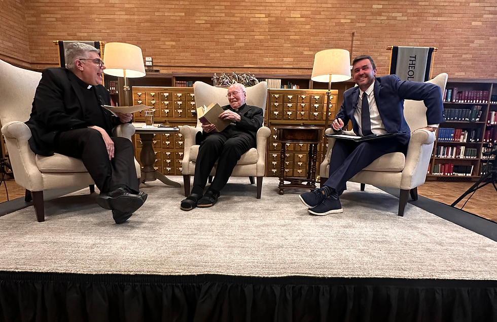 Three men sit on a stage in a library, engaged in conversation. Each holds a book or notebook. Warm lighting adds a cozy ambiance.