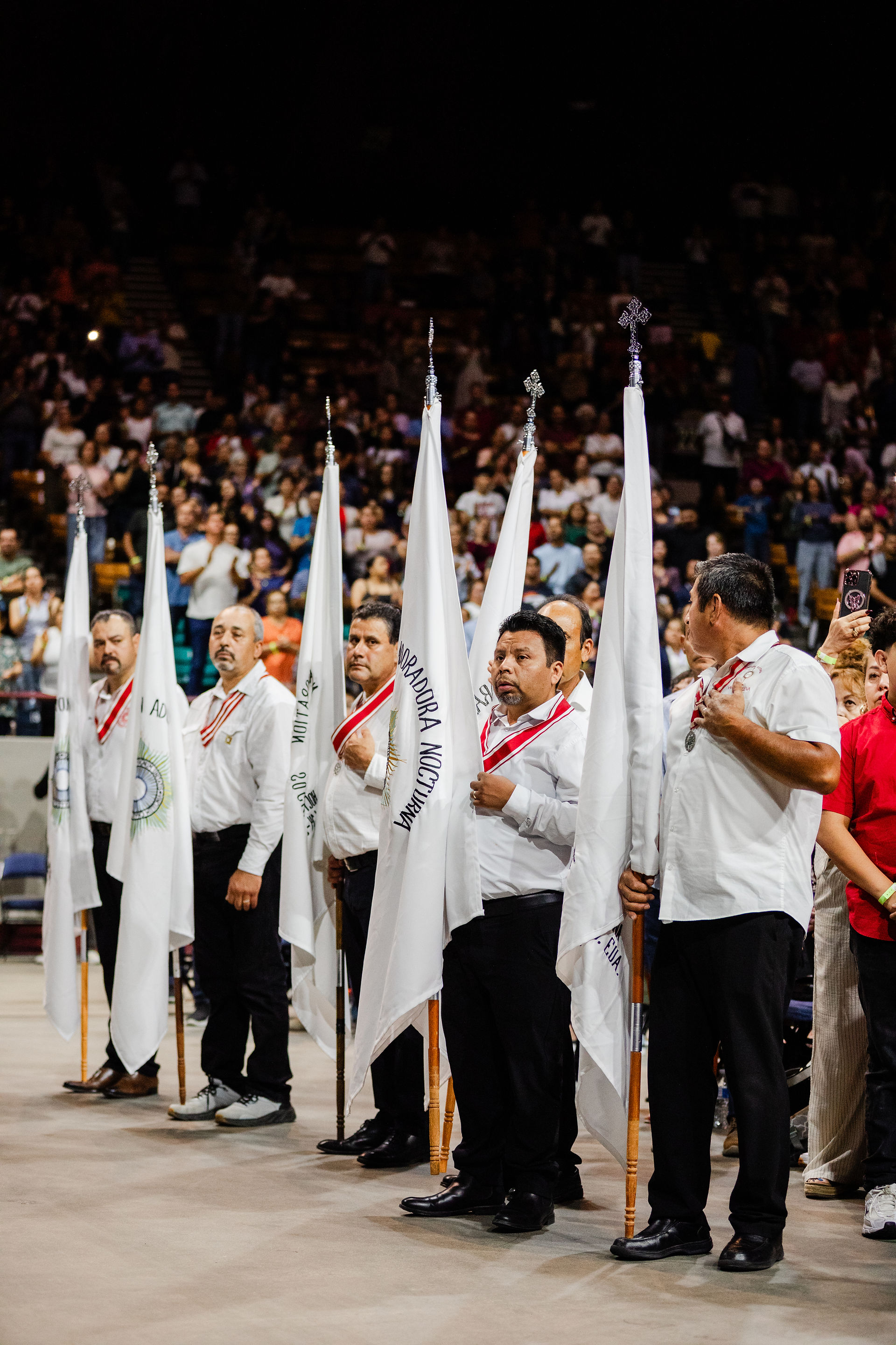 A line of men wearing white shirts and red medallions stands holding white flags with various Catholic group names written on them, during a large religious gathering. Behind them, a crowd of people fills the arena-style seating, suggesting a major event or celebration of faith.