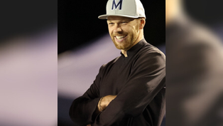 Smiling man in dark clothing with arms crossed, wearing a white cap with an "M" on it. Dark background creates a focused mood.