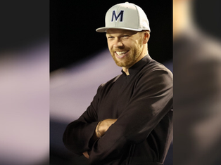 Smiling man in dark clothing with arms crossed, wearing a white cap with an "M" on it. Dark background creates a focused mood.