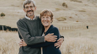 Older couple embracing, smiling in a grassy field with a fence and hills in the background. The mood is warm and joyful.