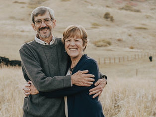 Older couple embracing, smiling in a grassy field with a fence and hills in the background. The mood is warm and joyful.