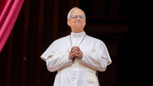 Man in white religious attire stands on a balcony, hands clasped, smiling. Red curtains in the background convey a formal setting.