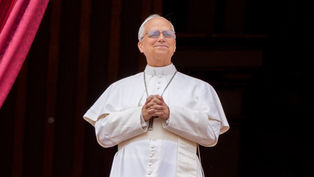 Man in white religious attire stands on a balcony, hands clasped, smiling. Red curtains in the background convey a formal setting.