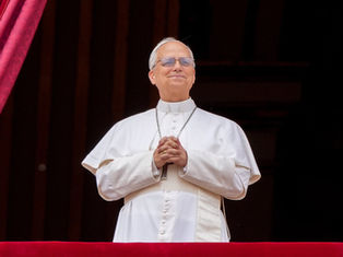 Man in white religious attire stands on a balcony, hands clasped, smiling. Red curtains in the background convey a formal setting.