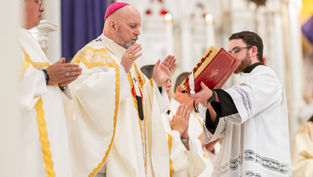 Clergy in white and gold robes during a religious service, one holding a red book. Ornate background, solemn mood in a church setting.