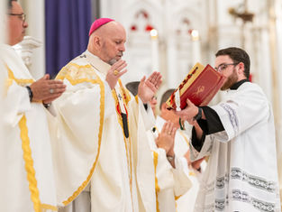 Clergy in white and gold robes during a religious service, one holding a red book. Ornate background, solemn mood in a church setting.