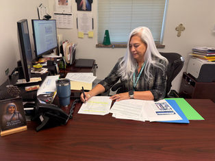 Woman with long white hair working at a desk, writing on documents. Office setting with computer, books, and a St. Teresa of Calcutta plaque.
