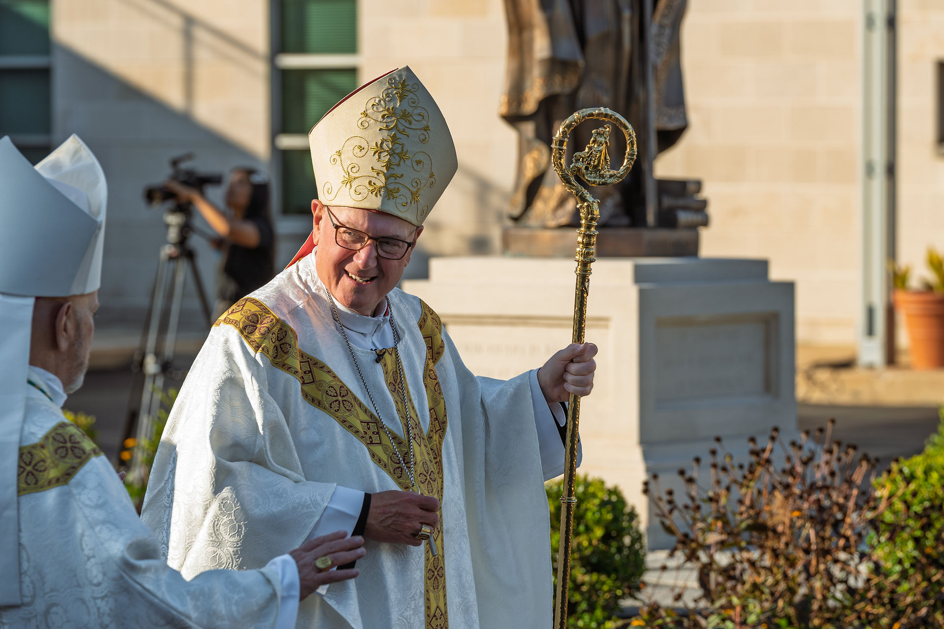 A smiling bishop in ornate robes and mitre holds a staff, conversing with another clergy. Background shows a statue and a camerawoman.