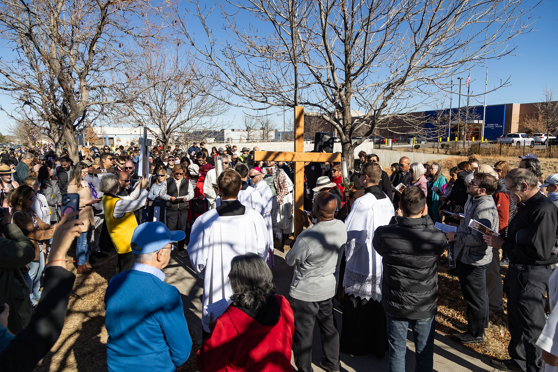 Crowd gathers for an outdoor religious procession with a large wooden cross. Clear sky, people in dark coats and white robes. Solemn mood.