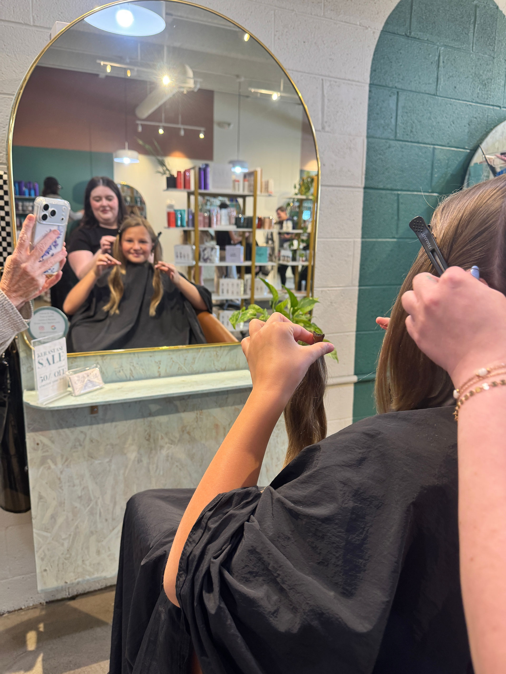 View in a mirror of a girl holding sections of her hair while someone prepares to cut it, with others watching and taking photos.