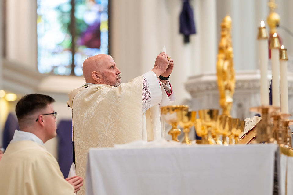 Priest in ivory robe raises a host during a church service. Gold chalices and candles are on the altar. Stained glass window in background.