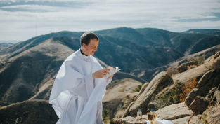 Priest in white robe reads a book on a rocky mountain, with a golden chalice and bottles nearby. Sunny, expansive landscape in background.