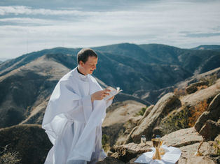 Priest in white robe reads a book on a rocky mountain, with a golden chalice and bottles nearby. Sunny, expansive landscape in background.