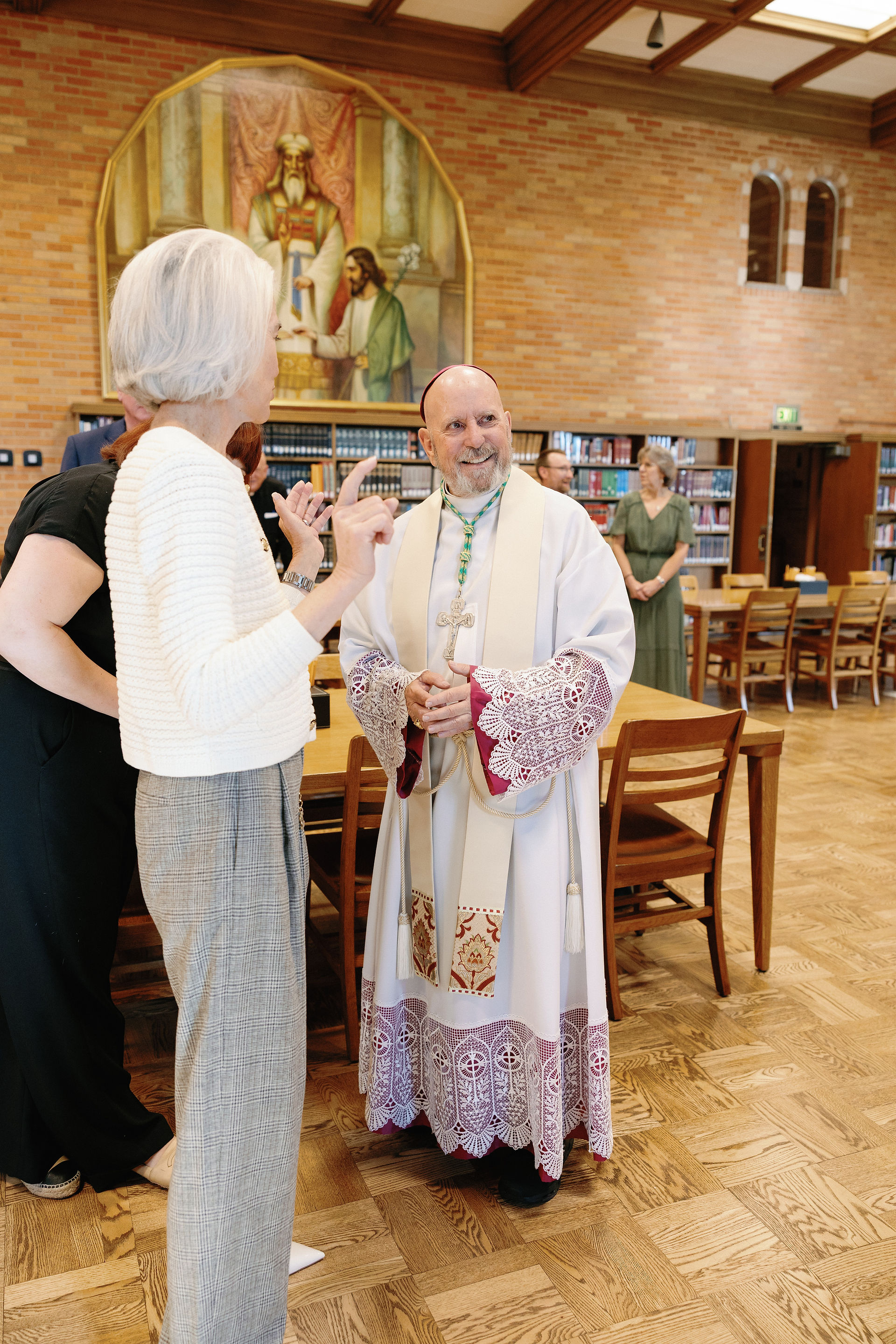 A bishop in ornate robes smiles while conversing with a woman in a library. Brick walls, a religious painting, and bookshelves in the background.