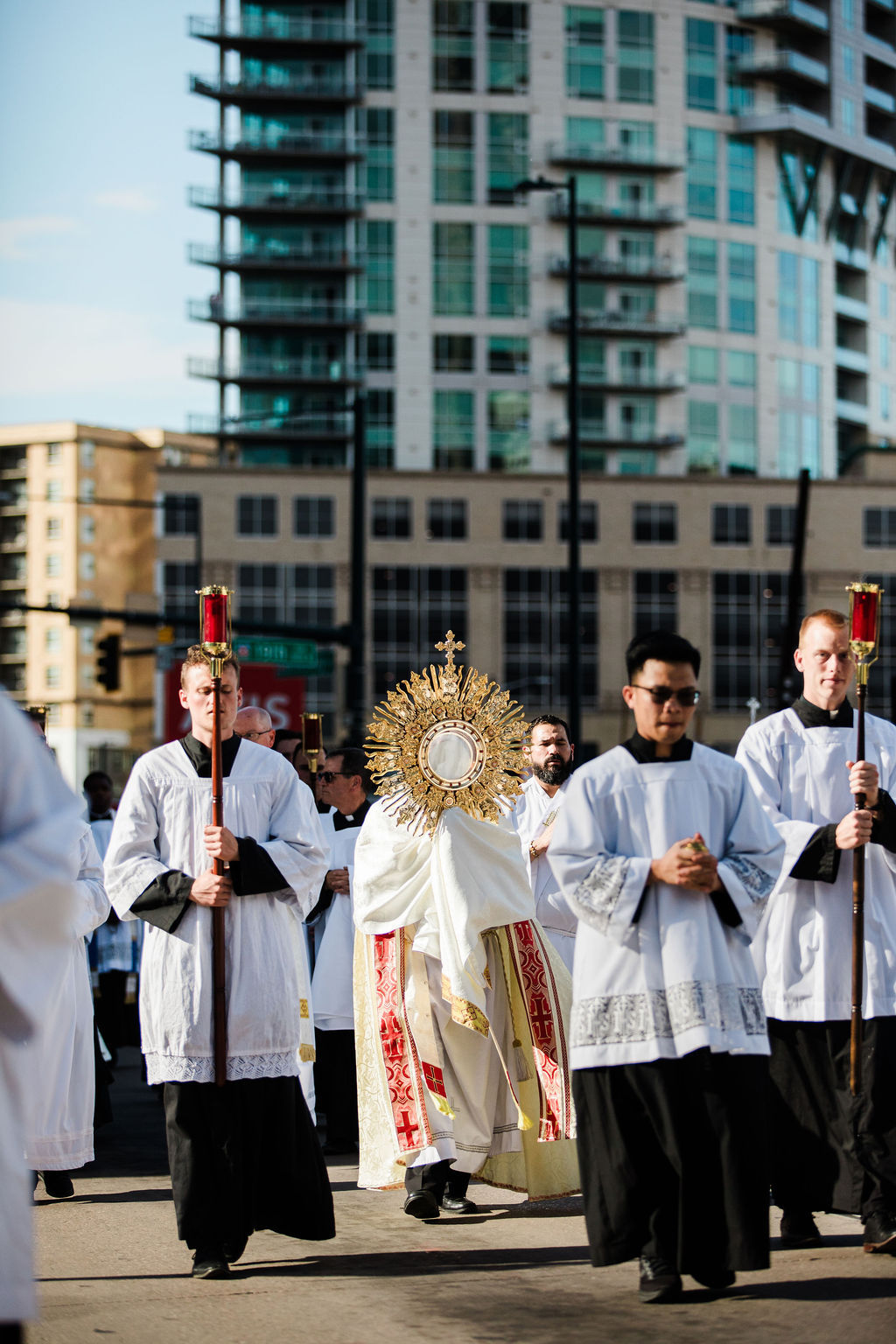 Catholic procession with priests in white robes carrying a golden monstrance and candles on a city street.