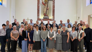 A group of 40 adults, some in religious attire, posing and smiling inside a church with stained glass and a statue in the background.