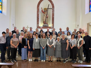A group of 40 adults, some in religious attire, posing and smiling inside a church with stained glass and a statue in the background.