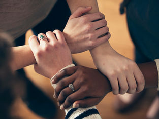 Diverse hands clasped together, forming a supportive circle on a wooden floor. Some wear rings; warm and cooperative mood.