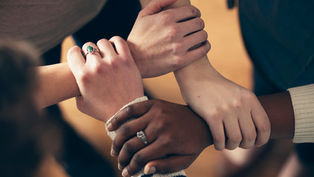 Diverse hands clasped together, forming a supportive circle on a wooden floor. Some wear rings; warm and cooperative mood.