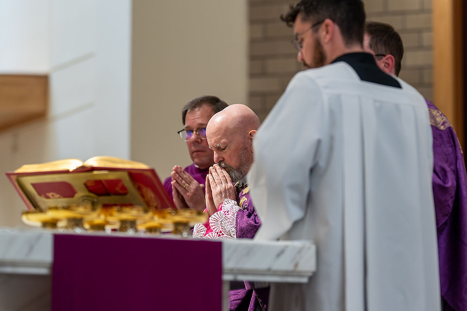 Bishop and clergy in purple robes pray at an altar with an open book and chalices. Sacred and reverent atmosphere.