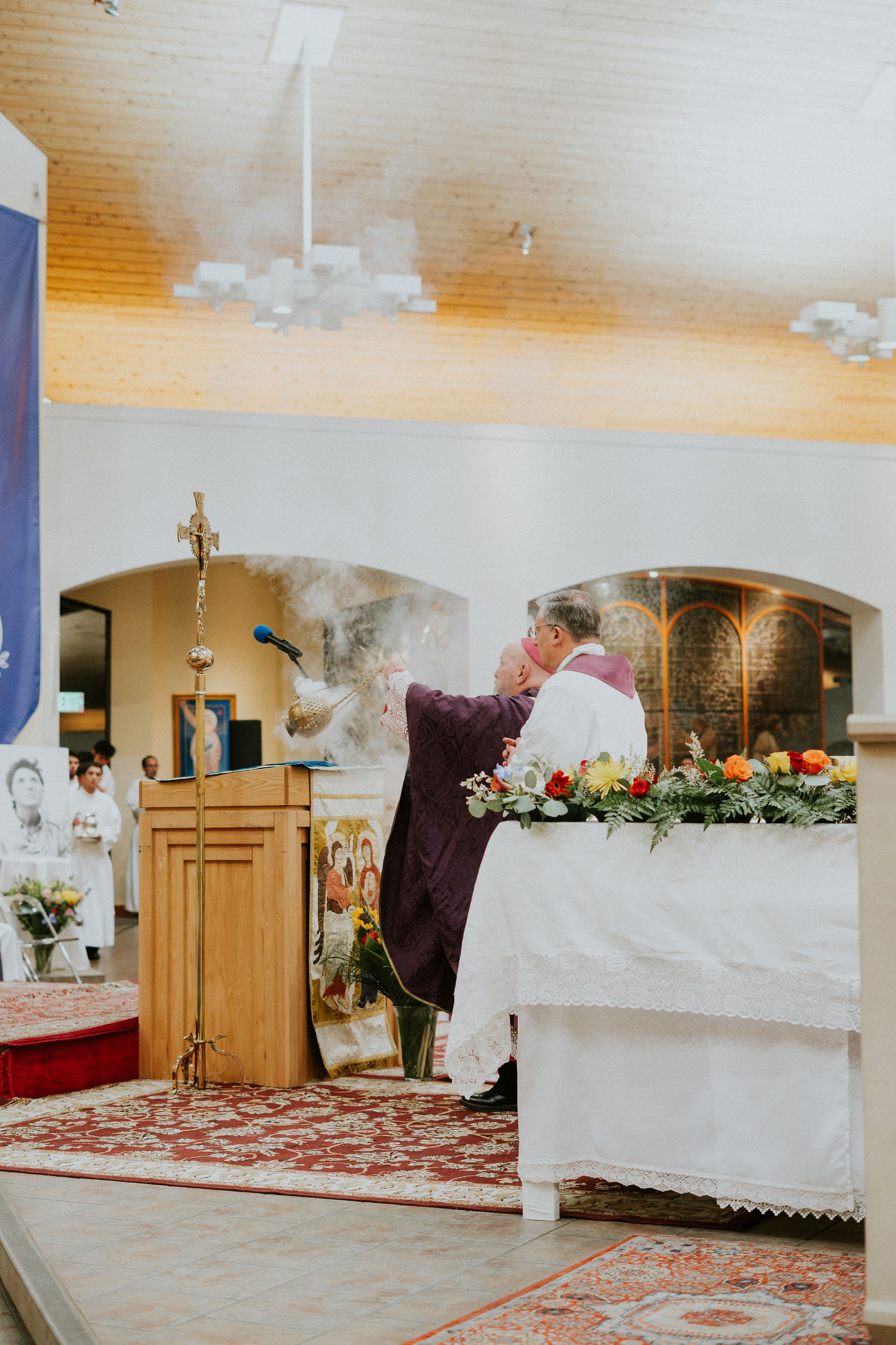 Clergy performing incense ritual in a church, with altar flowers and religious icons. Warm lighting and a patterned carpet enhance the setting.