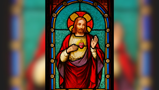 Stained glass of a haloed figure in red and white robes with a sacred heart on the chest, set against a turquoise background with ornate details.