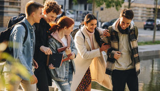 Five friends walking near a pond, laughing and carrying books. They wear casual fall attire. Background shows trees and buildings.