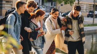 Five friends walking near a pond, laughing and carrying books. They wear casual fall attire. Background shows trees and buildings.