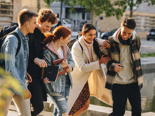 Five friends walking near a pond, laughing and carrying books. They wear casual fall attire. Background shows trees and buildings.