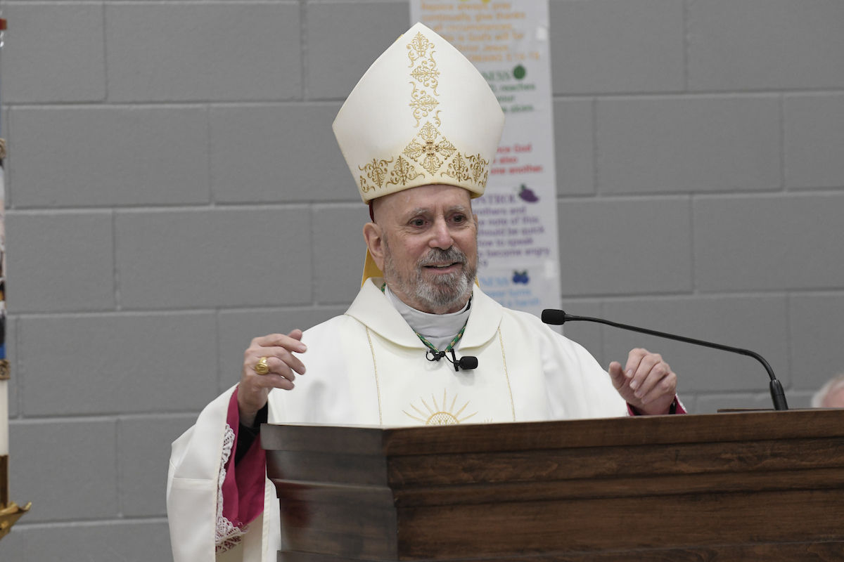 A bishop in ornate attire speaks at a wooden podium against a gray brick wall. A poster with text is partially visible in the background.