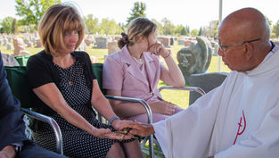 A somber outdoor gathering at a cemetery. A woman in a black polka-dot dress holds a priest's hand. Another woman in pink looks on.