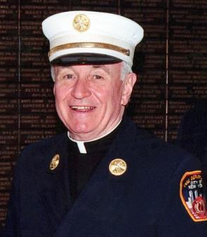 Smiling older man in a dark navy uniform and white cap stands before a dark plaque wall, with a fire/rescue patch on his sleeve.