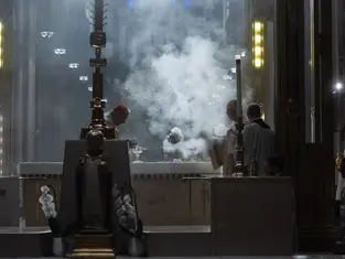 Clergy perform a ritual with incense smoke in a dimly lit cathedral. Ornate altar with white flowers; solemn and reverent atmosphere.