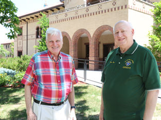 Two smiling older men stand outside a brick building, one in a red plaid shirt and one in a green polo.