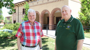 Two smiling older men stand outside a brick building, one in a red plaid shirt and one in a green polo.