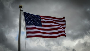 U.S. flag waving against a cloudy, gray sky. Red, white, and blue colors stand out, creating a solemn and proud atmosphere.