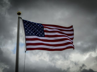 U.S. flag waving against a cloudy, gray sky. Red, white, and blue colors stand out, creating a solemn and proud atmosphere.