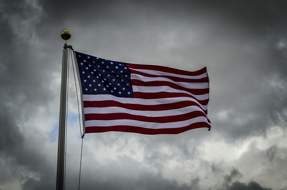 U.S. flag waving against a cloudy, gray sky. Red, white, and blue colors stand out, creating a solemn and proud atmosphere.