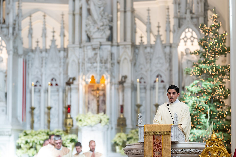 Deacon in white robe speaks at ornate pulpit in decorated church, with Christmas tree and candles in background; solemn atmosphere.