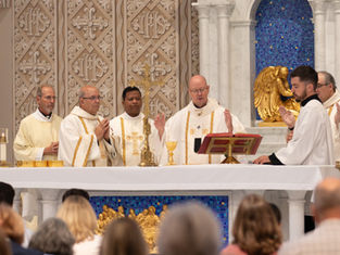Clergy in white vestments celebrate Mass at a gold altar before a blue mosaic backdrop, with worshippers seated in front.