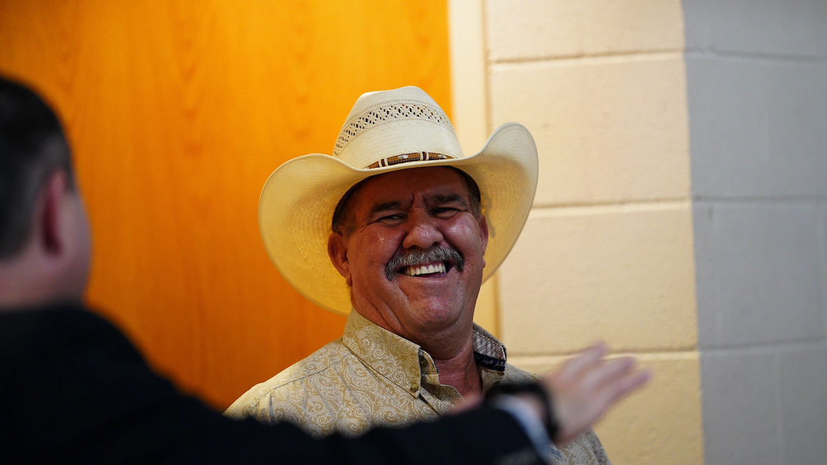Man in a cowboy hat smiles broadly, wearing a patterned shirt. Blurred foreground figure gestures. Background has light and wooden tones.