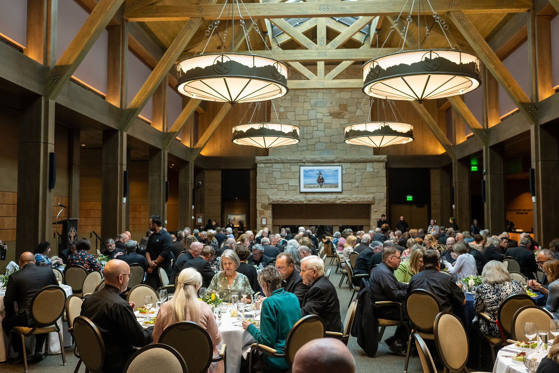 A large gathering in a wooden hall with round tables, chandeliers, and a stone wall. People are dining, chatting, and dressed formally.