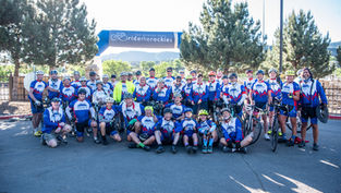 Cyclists in matching blue and white jerseys pose under a "Ride the Rockies" banner, smiling against a backdrop of trees and mountains.