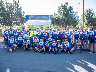 Cyclists in matching blue and white jerseys pose under a "Ride the Rockies" banner, smiling against a backdrop of trees and mountains.